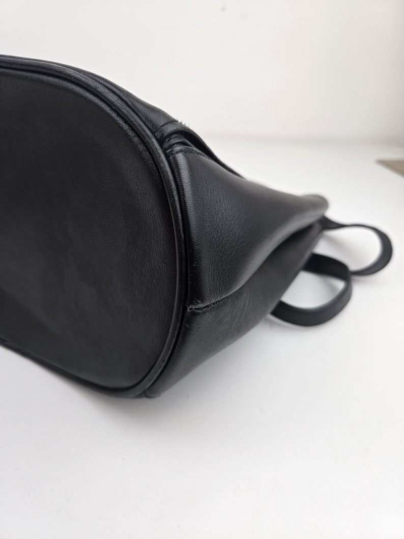 Pu&ograve; includere: Primo piano di una borsa in pelle nera. La borsa ha un fondo arrotondato e una consistenza liscia. Sono visibili due piccole maniglie. La borsa sembra vuota ed &egrave; posta su uno sfondo bianco.
