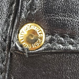 Pu&ograve; includere: Primo piano di pelle nera con un bottone dorato. Il bottone &egrave; rotondo e presenta la scritta "HIGH QUALITY LEATHER" incisa sul bordo. La pelle ha cuciture visibili.