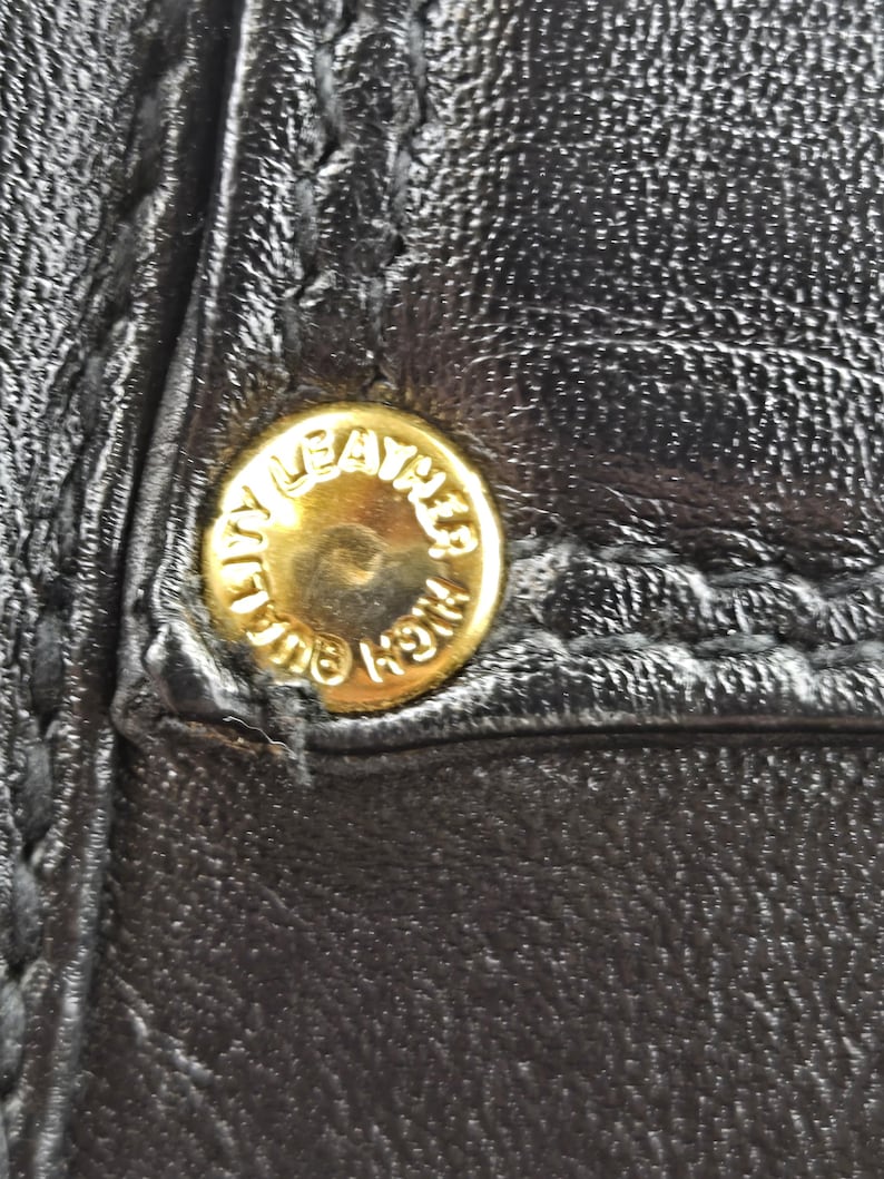 Pu&ograve; includere: Primo piano di pelle nera con un bottone dorato. Il bottone &egrave; rotondo e presenta la scritta "HIGH QUALITY LEATHER" incisa sul bordo. La pelle ha cuciture visibili.