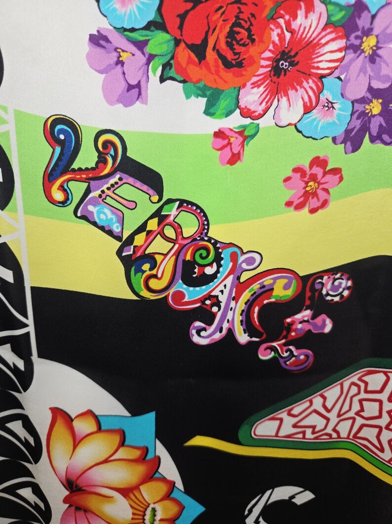 Puede incluir: Una tela con un estampado floral vibrante que presenta rosas rojas, flores moradas y azules, y hojas verdes. La palabra VERSACE est&aacute; impresa en una fuente estilizada y multicolor sobre un fondo de rayas horizontales verde lima, amarillo y negro.