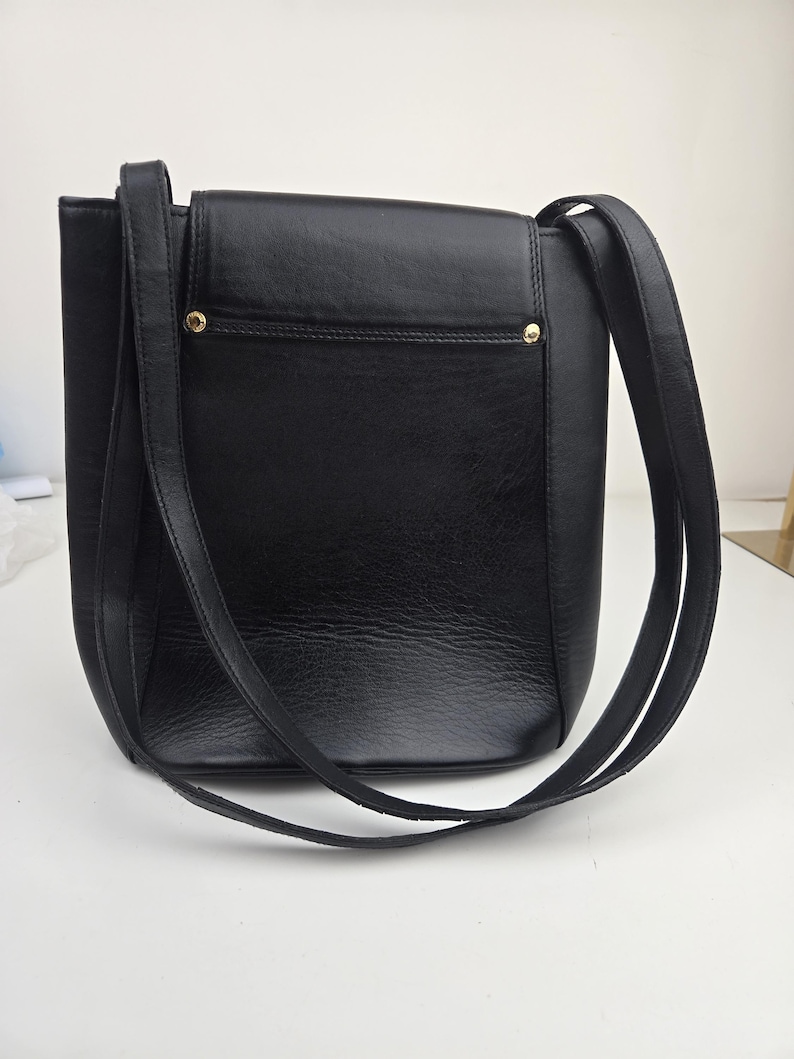 Pu&ograve; includere: Borsa a tracolla in pelle nera con chiusura a patta e lunghe cinghie. La borsa ha una forma rettangolare con una superficie strutturata e dettagli dorati. Le cinghie sono sottili e sembrano essere realizzate nello stesso materiale della borsa.