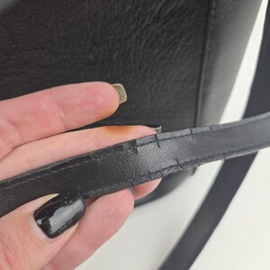 Pu&ograve; includere: Primo piano di una borsa in pelle nera con tracolla. La tracolla mostra segni di usura, con crepe visibili sulla superficie. &Egrave; visibile anche il corpo in pelle strutturata della borsa.