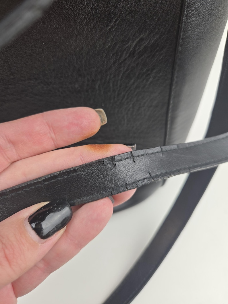Pu&ograve; includere: Primo piano di una borsa in pelle nera con tracolla. La tracolla mostra segni di usura, con crepe visibili sulla superficie. &Egrave; visibile anche il corpo in pelle strutturata della borsa.