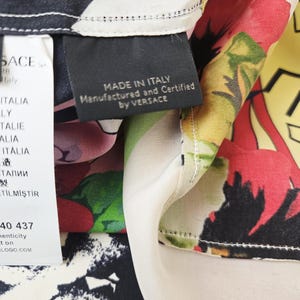 Puede incluir: Una tela de seda con un vibrante estampado floral en rojo, verde, amarillo y morado sobre un fondo negro y crema. Una etiqueta negra dice MADE IN ITALY Manufactured and Certified by VERSACE, junto a una etiqueta blanca con VERSACE y texto.