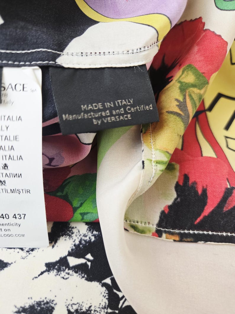 Puede incluir: Una tela de seda con un vibrante estampado floral en rojo, verde, amarillo y morado sobre un fondo negro y crema. Una etiqueta negra dice MADE IN ITALY Manufactured and Certified by VERSACE, junto a una etiqueta blanca con VERSACE y texto.