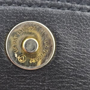 Pu&ograve; includere: Primo piano di un articolo in pelle nera con chiusura magnetica argentata. La chiusura riporta le scritte "UK PAT 1519246" e "UK D973682". La pelle presenta cuciture visibili.