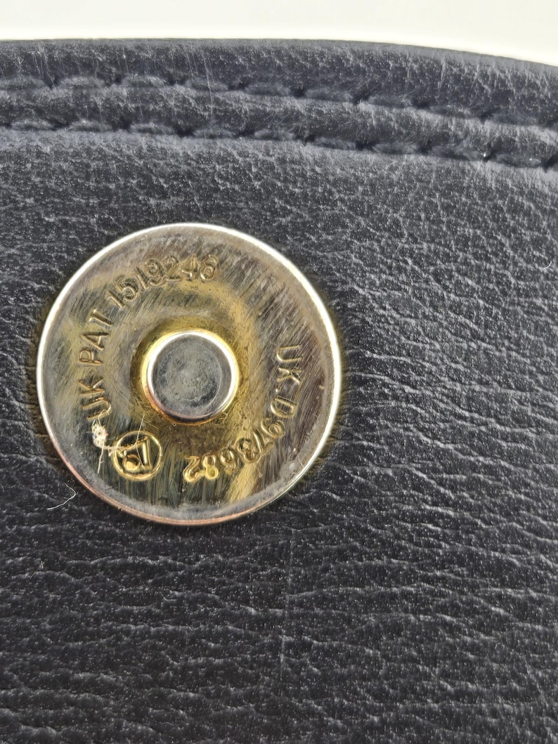 Pu&ograve; includere: Primo piano di un articolo in pelle nera con chiusura magnetica argentata. La chiusura riporta le scritte "UK PAT 1519246" e "UK D973682". La pelle presenta cuciture visibili.