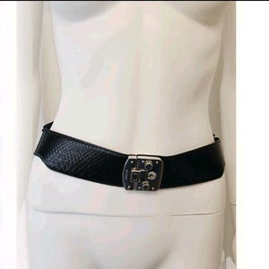 Puede incluir: Un cinturón negro con un patrón texturizado de piel de serpiente y una hebilla de metal plateado. La hebilla tiene una forma rectangular con un mecanismo de cierre central y la palabra Gucci grabada en el lado izquierdo del componente.