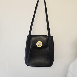 Pu&ograve; includere: Borsa a tracolla in pelle nera con una lunga tracolla. La borsa ha una chiusura a patta con un emblema circolare dorato. La borsa &egrave; appesa a una parete bianca.