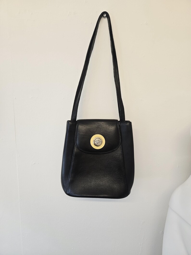 Pu&ograve; includere: Borsa a tracolla in pelle nera con una lunga tracolla. La borsa ha una chiusura a patta con un emblema circolare dorato. La borsa &egrave; appesa a una parete bianca.