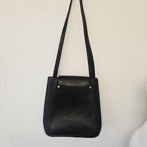 Pu&ograve; includere: Una borsa a tracolla in pelle nera con una lunga tracolla, appesa a una parete bianca. La borsa ha una forma rettangolare con chiusura a patta e dettagli color oro. La pelle ha un aspetto strutturato.