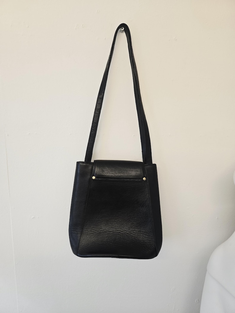 Pu&ograve; includere: Una borsa a tracolla in pelle nera con una lunga tracolla, appesa a una parete bianca. La borsa ha una forma rettangolare con chiusura a patta e dettagli color oro. La pelle ha un aspetto strutturato.