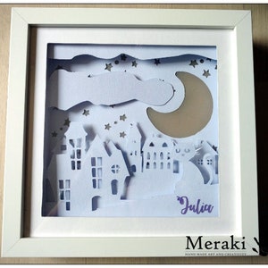Puede incluir: Arte de papel recortado blanco en un marco blanco. El arte representa una escena nocturna con una luna creciente, estrellas, nubes y casas. El nombre "Julia" está escrito en morado en la parte inferior del arte.