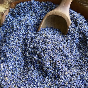 May include: A large quantity of dried purple lavender buds filling a wooden bowl, with a smooth wooden scoop resting partially submerged in the center of the fragrant floral pile, showcasing the deep blue and violet tones of the dried plant material.