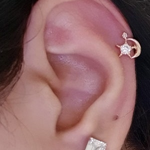 May include: A close-up of an ear with a gold-colored earring featuring a star and crescent moon design, adorned with clear gemstones. A second, rectangular gemstone earring is also visible.