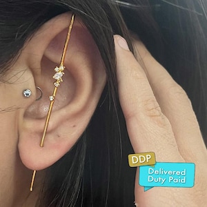 Könnte beinhalten: Ein goldener Ohrklemme mit linearem Design, verziert mit kleinen, funkelnden Edelsteinen. Die Manschette wird am Ohr getragen und zeigt ihre zarte und elegante Ästhetik. Das Ohr hat auch einen kleinen silbernen Ohrstecker und einen gebogenen Barbellen-Ohrring.