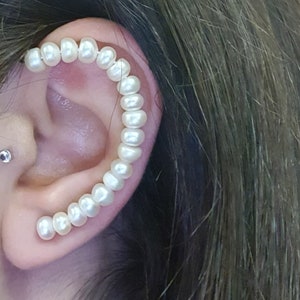 May include: A curved ear cuff featuring a series of white pearls. The pearls are strung together to create a decorative design that follows the curve of the ear. The jewelry is a delicate and elegant accessory.