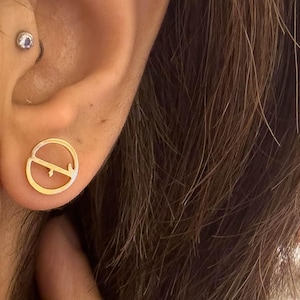 May include: Gold-colored stud earrings with a peace symbol design. The earrings are circular with a thin outline and a small, straight line through the center. The earrings are worn on a person's ear.