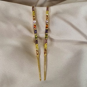 May include: A pair of gold-coloured earrings with a linear design. Each earring features a series of colourful, rectangular gemstones in shades of yellow, green, purple, orange, and pink. The earrings have a long, slender, dangling style.