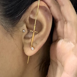 May include: A gold-colored ear cuff featuring a sword design. The sword has a cross-shaped hilt with a red gem and a small, clear gem at the tip. The earring is worn on the ear.