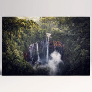 Könnte beinhalten: Ein Foto eines Wasserfalls, der durch einen dichten Wald stürzt. Das Wasser fällt eine Klippe hinunter und erzeugt Nebel am Boden. Das Bild ist ein Landschaftsdruck mit grünen Bäumen und einer natürlichen Szene.