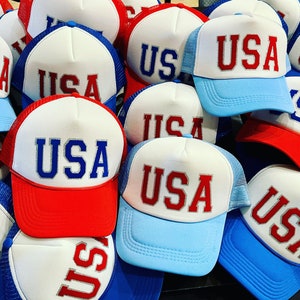 May include: A pile of red, white, and blue trucker hats with the word "USA" embroidered on the front.
