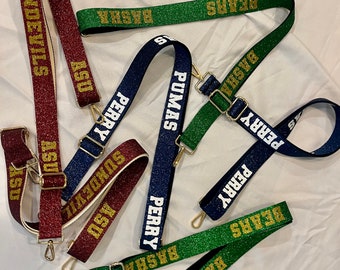 May include: A collection of colorful, glittery straps with gold hardware and various school names printed on them, including "ASU", "SUNDEVILS", "PERRY", "PUMAS", "BASHA", and "BEARS".