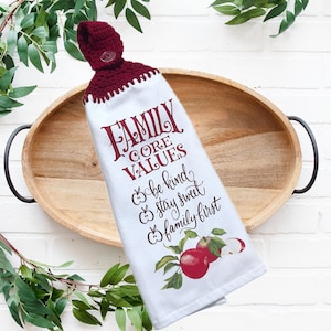 May include: White kitchen towel with a burgundy crocheted top and button. The towel features the text "FAMILY CORE VALUES" and the phrases "be kind, stay sweet, family first" with apple illustrations. The towel is on a wooden tray.