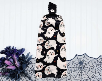 May include: A black and white Halloween kitchen towel with a loop for hanging. The towel features a pattern of white cartoon ghosts with pink cheeks and orange stars on a black background. The top loop is black and has a white button.