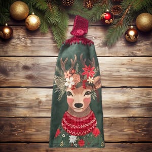 May include: A dark green hanging kitchen towel featuring a central illustration of a deer with a floral crown and a red plaid scarf. The top of the towel has a maroon crocheted topper with a matching button for hanging on a cabinet handle.