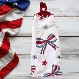 May include: A white kitchen towel with a red, white, and blue bow and stars design. The towel has a red crocheted loop with a button at the top.