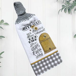May include: White kitchen towel with a gray crocheted top and a gingham bottom border. The towel features a bee-themed design with a beehive illustration and the words "HIVE RULES" and "BEE POSITIVE".