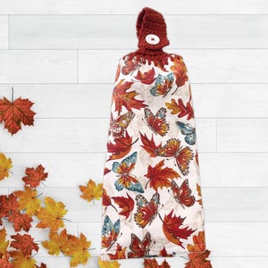 May include: A hanging kitchen towel with a burgundy crocheted top and a white button closure. The towel features a pattern of colorful butterflies and autumn leaves in shades of red, orange, and blue on a white background.