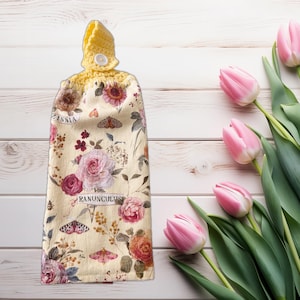 May include: A hanging kitchen towel with a floral design featuring roses, butterflies, and the words "Zinnia" and "Ranunculus." The towel has a yellow crocheted top with a button closure. Pink tulips are in the background.