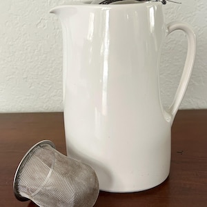 May include: A white ceramic jug with a curved handle and spout, alongside a stainless steel tea infuser. The infuser is a mesh basket design. The jug is on a wooden surface.