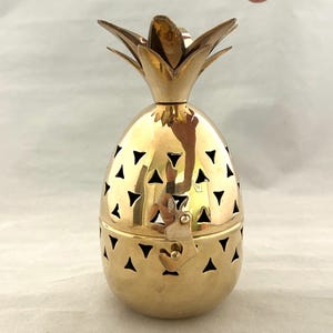Op de afbeelding: Een gepolijste goudkleurige metalen houder in de vorm van een ananas met een afneembaar deksel. Het lichaam is voorzien van een herhalend patroon van driehoekige uitsparingen en een klein metalen sluitmechanisme aan de voorzijde voor de helften.