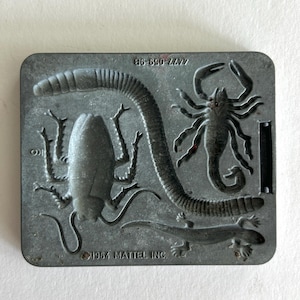 May include: A vintage metal mold featuring insect and reptile shapes. The mold includes a worm, cockroach, scorpion, and lizard. The text "1964 Mattel Inc." is visible. The mold is rectangular with rounded corners and a dark gray color.