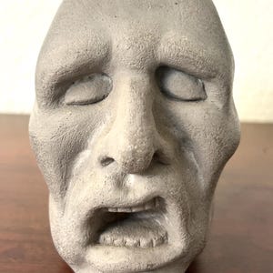 May include: A gray, textured sculpture of a human head featuring closed eyes, a prominent nose, and an open mouth. The surface has a matte, stone-like appearance with visible fine lines and contours that define the facial features and expression.