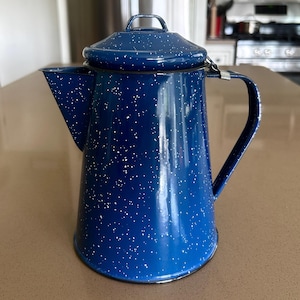 May include: A deep blue enamel coffee percolator featuring a white speckled pattern throughout the exterior. The vessel includes a matching lid with a small handle, a wide pouring spout, and a sturdy side handle attached with a metal bracket.