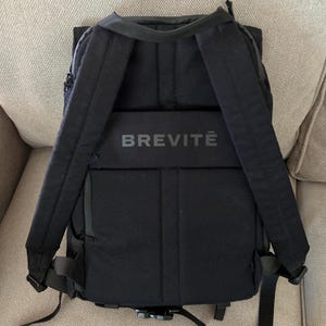 Puede incluir: Una mochila negra con correas acolchadas para los hombros y una cinta horizontal para equipaje en el panel posterior. La palabra BREVITĒ está impresa en color gris oscuro en la cinta horizontal central de esta bolsa negra estructurada.