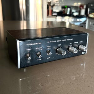 May include: A vintage Realistic SA-10 solid state stereo amplifier with a black front panel, silver knobs, and toggle switches. The unit features labels for power, selector, mode, tone, and volume controls, housed in a dark wood-grain case.