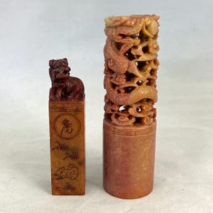 May include: Two carved stone seals. The left seal is rectangular with a dark brown lion figure on top, featuring an engraved circle with Chinese characters and a small animal motif. The right seal is cylindrical with an intricate dragon carving.