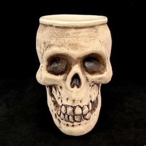May include: A ceramic skull-shaped container with a wide opening. The skull is off-white with dark eye sockets and a detailed jawline. The container is likely used for decorative or novelty purposes.