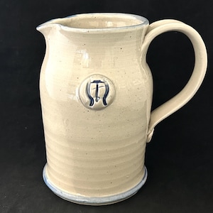 May include: A beige ceramic jug with a spout and large handle. The jug has a blue rim and a blue emblem on the front. The jug is approximately 23 cm tall.