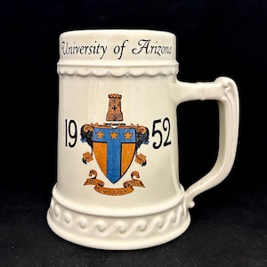 May include: Cream-colored ceramic beer stein with a handle, featuring the words "University of Arizona" and the year "1952." The stein has a decorative crest with a tower, stars, and the Greek letters "Pi" and "Upsilon."