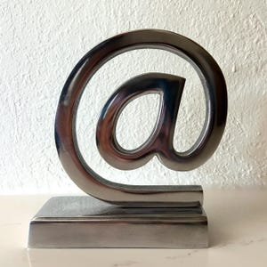 Op de afbeelding: Een sculptuur van gepolijst zilverkleurig metaal in de vorm van het apenstaartje. Het ontwerp bestaat uit een doorlopende gebogen lijn die het teken vormt, stevig gemonteerd op een massieve, rechthoekige metalen voet met een gladde, reflecterende afwerking.