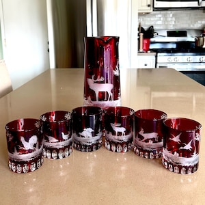 Puede incluir: Un jarro de vidrio rojo rubí y seis vasos a juego. Cada pieza presenta un diseño blanco grabado de fauna, incluyendo ciervos y aves. La cristalería se exhibe sobre una encimera de color claro.