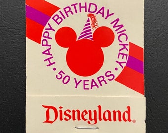 Peut inclure: Un livre d'allumettes blanc avec un motif rouge et violet. Le devant présente une tête de Mickey Mouse rouge portant un chapeau de fête rayé violet et blanc. Le texte indique "Happy Birthday Mickey 50 Years". Le dos du livre d'allumettes indique "Disneyland".