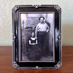 May include: A rectangular picture frame with a glossy black enamel finish and silver-toned metal trim. The frame features intricate scrollwork at the corners and edges, accented with small clear crystals. It holds a black and white photograph.
