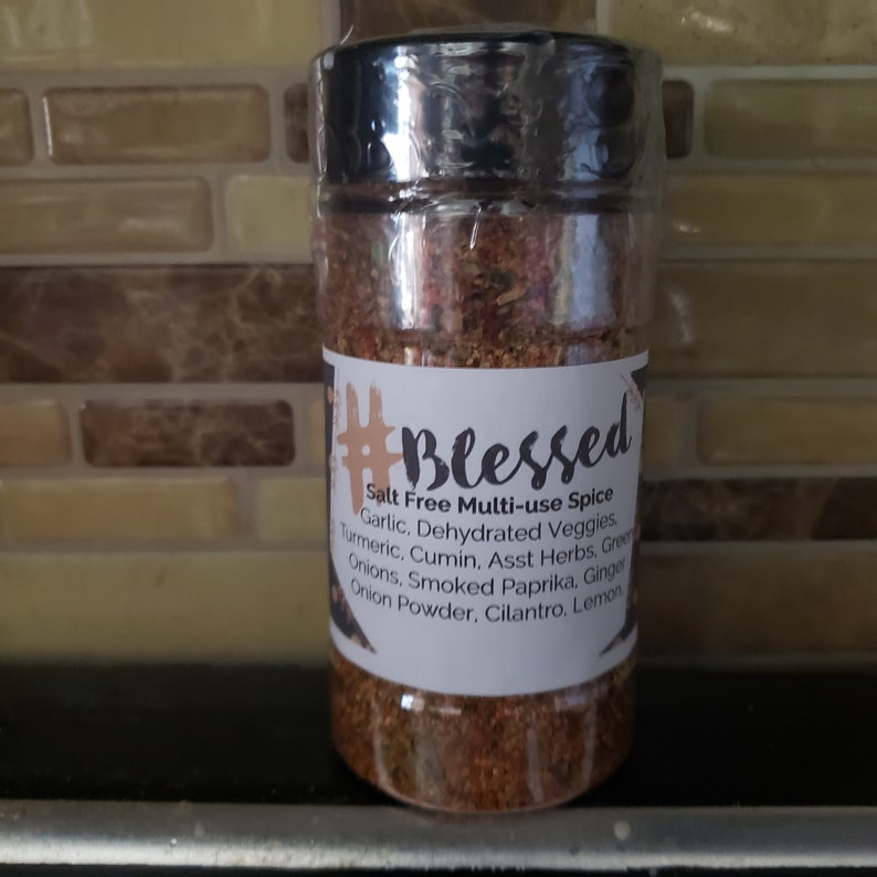 May include: A glass jar filled with a blend of spices labelled "#Blessed Salt Free Multi-use Spice". The label lists the ingredients: Garlic, Dehydrated Veggies, Turmeric, Cumin, Asst Herbs, Green Onions, Smoked Paprika, Ginger, Onion Powder, Cilantro, Lemon.